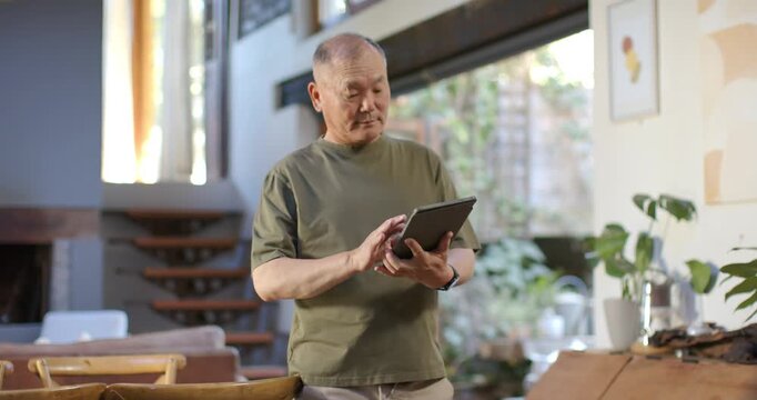 Senior Asian man in living room holding tablet, tapping, swiping screen, reading and glancing right