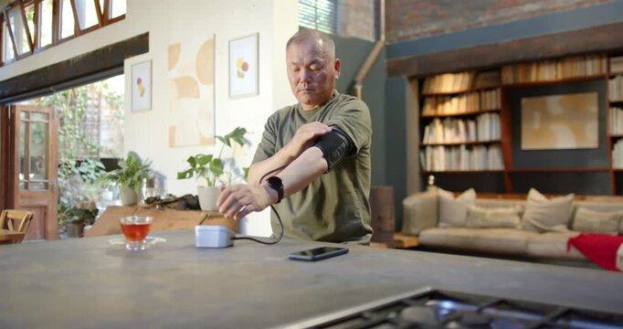 Senior Asian man fitting cuff, pressing monitor on kitchen island, checking blood pressure reading