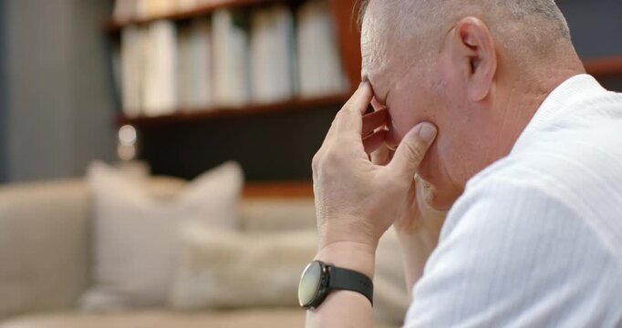 Senior Asian man sitting right sofa, pinching nose and rubbing eyes from strain, watch, copy space