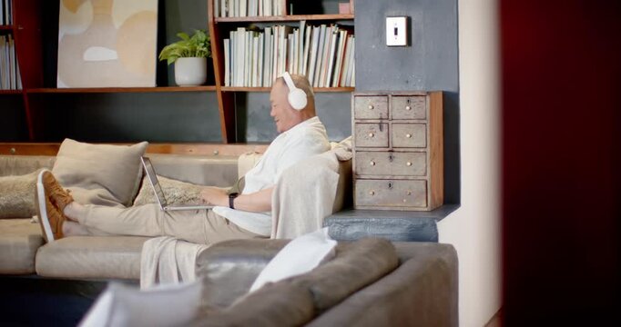 Camera clearing Sr Asian man lounging on couch typing leisurely on laptop with phones, copy space