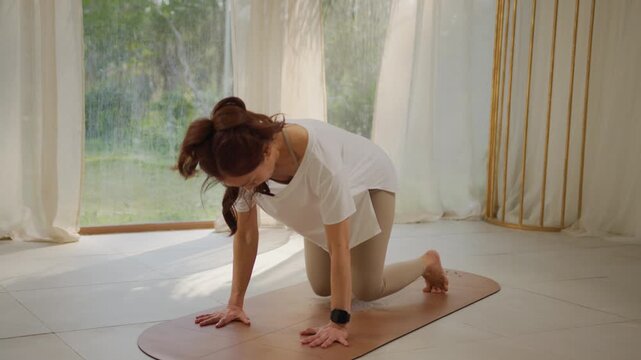Woman yoga indoor exercise stretch pose on mat in bright home room with soft window light, calm fitness practice, flexible body movement and serene morning workout