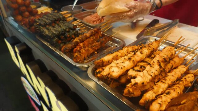 Street food vendor places a bread roll on a board beside trays of grilled skewers, wrapped meats and fried snacks at a busy asian night market stall, ready to sell