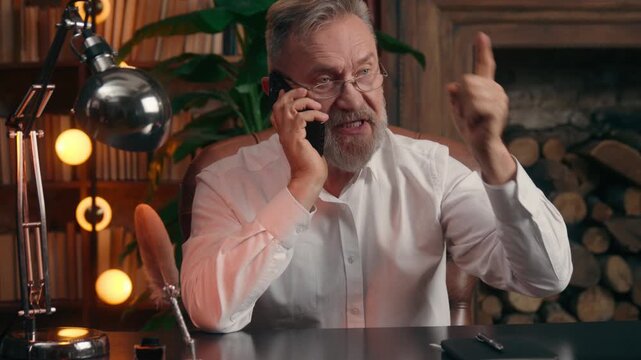 Negative irritated Caucasian old retired business man arguing mobile phone angry talking call conflict quarrel with client work problem at office table annoyed senior aged businessman male argue talk