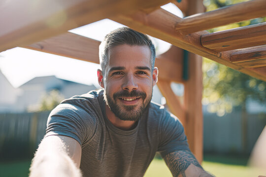 A smiling man with a beard enjoys outdoor exercise on wooden monkey bars in a sunny backyard.