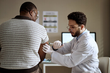 Medium shot of Black middle aged man receiving insulin injection from Caucasian male doctor during clinic visit. Supporting immunization, preventive healthcare, medical campaign use © Seventyfour