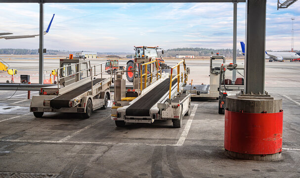 Belt loaders and ground support equipment stand ready on the airport tarmac with an airplane visible in the background.