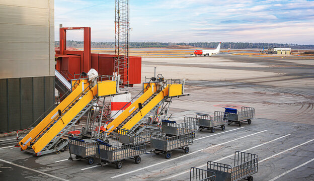 Ground support equipment including yellow passenger stairs and baggage carts sit on an airport apron with an airplane taxiing in the background under a soft sunset sky.