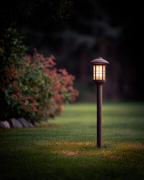A lantern-style garden light softly illuminates a green lawn at dusk, bordered by lush bushes and flowers.