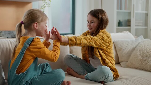 Two energetic Caucasian girls siblings playing patty cake game together on sofa European joyful children clapping hands entertainment smiling kids sisters play in living room clap palms at home couch