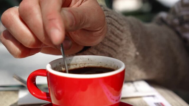 Hand stirs freshly served black coffee in a bright red cup at an outdoor cafe table. Concept of morning ritual, cozy break, and urban lifestyle
