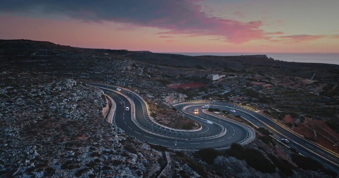 Malta: Aerial view of cars driving on a winding road on the coast of Malta at bright sunset, creating a scenic landscape with the Mediterranean Sea in the background. Drone flight
