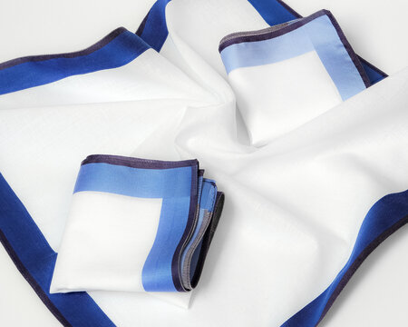 Two folded white cotton handkerchiefs with blue silk borders placed on an unfolded handkerchief with deep blue edges.