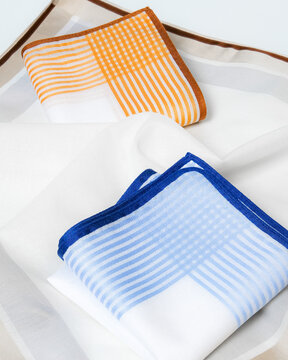 Elegant composition of two folded blue and orange striped handkerchiefs placed on an unfolded white cotton handkerchief with beige borders.