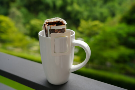 Single serve drip coffee filter bag brewing in a white ceramic mug on balcony railing with lush green blurred nature background. Morning travel coffee concept, portable fresh brew outdoors.
