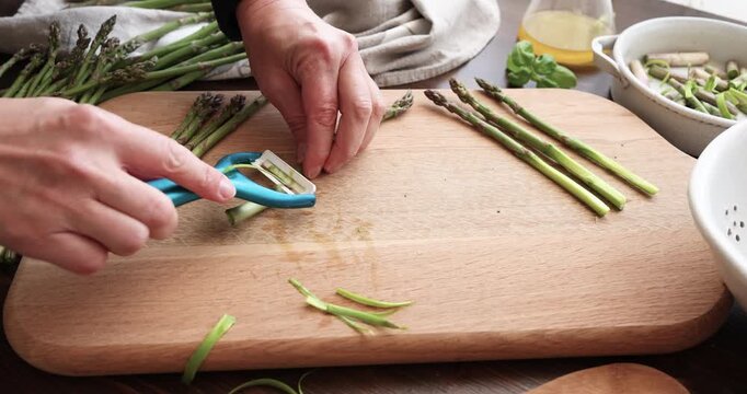 Woman hands peeling fresh green asparagus with blue vegetable peeler on wooden board. Chef preparing organic ingredients for healthy meal in domestic kitchen. Process of cleaning raw vegetables for