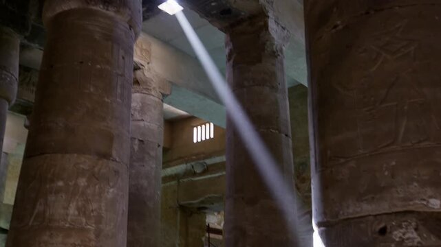 Cinematic interior of Abydos temple, Egypt, with powerful shaft of natural light breaking through window into massive dark colonnade of carved sandstone columns.