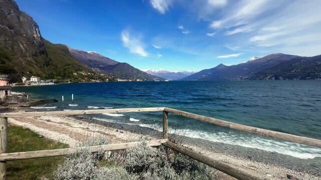 Lake como shoreline with waves pebbled beach wooden fence and mountain panorama under blue sky scenic italian landscape travel destination