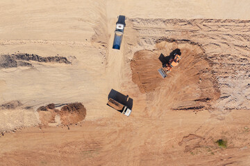 Construction site with heavy machinery, including an excavator loading soil into multiple dump trucks, performing large-scale earthmoving and development work from an aerial perspective © Nasos Zovoilis