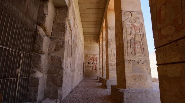 Colonnade corridor inside Abydos temple of Seti I, Egypt, with carved hieroglyphic reliefs on walls, square columns and vivid painted pharaoh figure visible at the illuminated far end.
