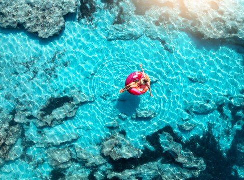 Aerial drone view of woman relaxing on pink inflatable ring in clear turquoise sea above rocks at sunset. Summer vacation, Mallorca, Balearic Islands, Spain. Top down view of swimming girl, blue water