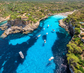 Aerial drone view of yachts and boats floating in narrow turquoise sea bay with rocky cliffs, green trees, sandy beach and swimmers, summer vacation scene. Mallorca, Balearic Islands, Spain. Top view © den-belitsky