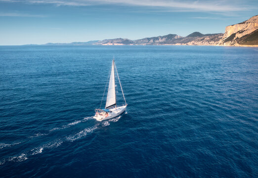 Aerial view of floating sailboat and rocky sea coast on summer sunny day. Top drone view of yacht, mountains, blue water. Yachting in Sardinia, Italy. Tropical seascape with boat. Summer vacation