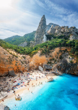 Aerial view of rocks and mountains, blue bay, sandy beach, people on sunny summer day. Cala Goloritze, Sardinia Island, Italy. Top drone view of cliffs, stones, green trees, transparent azure water