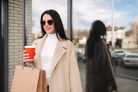 Woman in beige coat holding coffee and shopping bags with sunglasses by shop window reflection