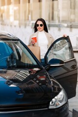 Obraz premium Stylish woman with coffee and shopping bags getting into a black car in the city