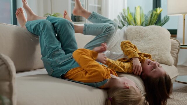 Two happy Caucasian little girls sisters lying upside down tickling laughing smiling kids children siblings friends daughters lie on couch sofa tickle play laugh smile fun energetic playing at home