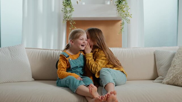 Two little Caucasian girls siblings kids children sisters friends daughters sharing secret tell gossip whispering on ear amazing news gossiping whisper friendly wondering on couch living room at home
