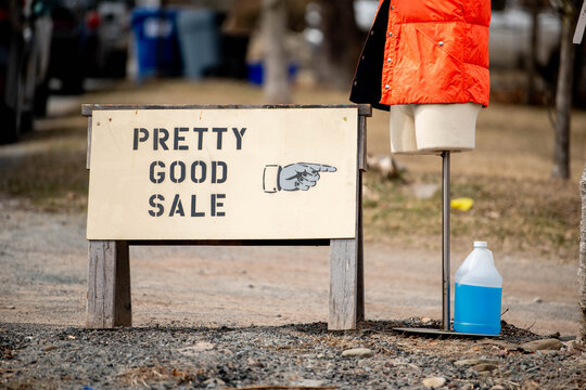  A handmade wooden sign reading "Pretty Good Sale" sits at a yard sale with an orange item hanging above it and a blue cleaning product beside it, a dryly funny piece of honest homemade advertising.