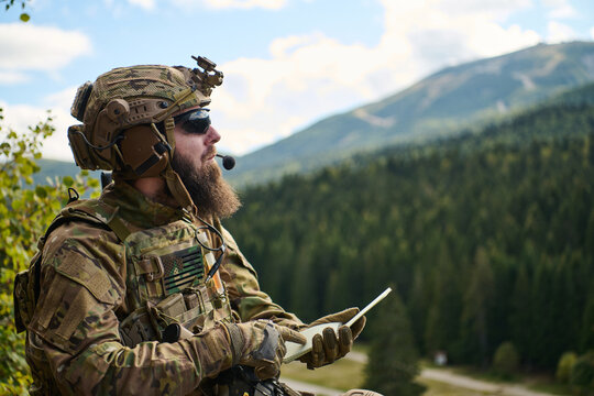 Bearded Soldier In Tactical Gear Using Tablet With Radio Headset And Mountain Landscape Background