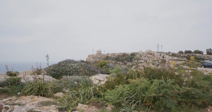Coastal radar station on a rugged cliff overlooking the calm sea with green vegetation under an overcast sky
