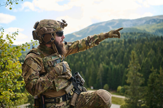 Bearded Soldier in Tactical Gear Pointing Toward Distant Mountains During Forest Reconnaissance Patrol
