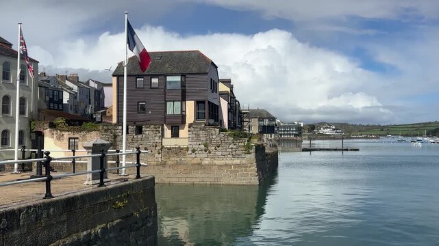 Static wide angle shot of Cornish and French flags gently waving above historic waterside buildings along the serene River Fal in Falmouth, Cornwall, England. Bright daylight, clear reflections.
