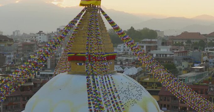 Drone shot of Boudhanath Stupa in Nepal with prayer flags and birds, set against warm sunset light over hills and city buildings.