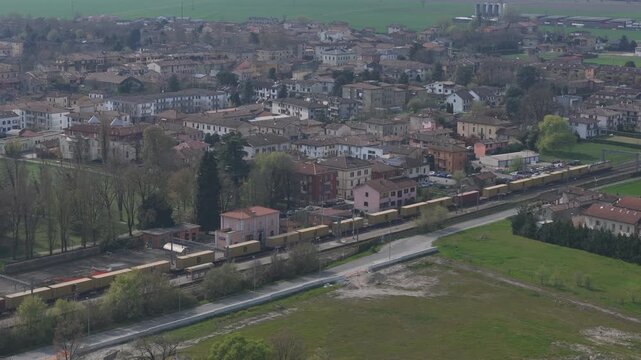 Freight train on Milano Bologna railway line passing Pontenure Piacenza Emilia Romagna Italy with urban settlement and surrounding Po Valley farmland, drone elevated lateral tracking shot