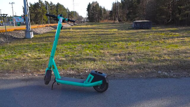 Greenish rental electric scooter parked on an asphalt path near a railway on a sunny day. Micromobility sharing services represent an eco friendly transport alternative for modern urban life