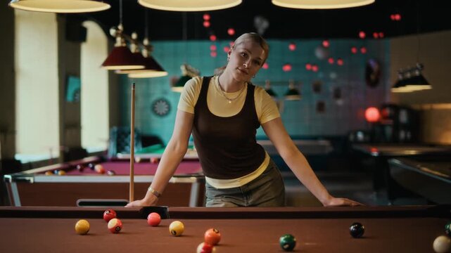 Dolly in portrait shot of smiling young adult woman facing camera while leaning on pool table during match at pool hall, slow motion