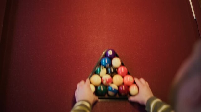 Top view of unrecognizable man arranging pool balls in triangle on red felt table using rack before beginning of game