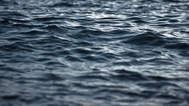 Close up detail of dark blue water with small waves and subtle reflections, showcasing a tranquil and rhythmic movement of fluid surface in low light conditions. Background slow motion. Close up