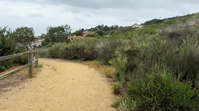 Unpaved hiking route, earthy tone, stretches through dense scrubland. Left side showcases weathered split rail fence. Right side features varied, drought tolerant plant life. Elevated background revea