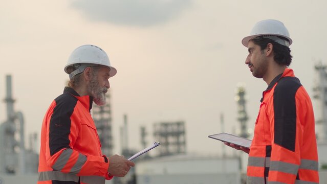 Two engineers collaborating on site management at an oil refinery, using a tablet and clipboard to review data for a quality control inspection at dusk.