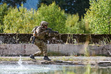 Naklejka premium Soldier In Camouflage With Rifle Running Along Water Splash During Military Training Exercise