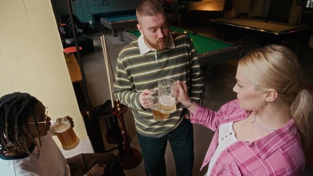 Pull out high angle shot of group of happy young adults clinking beers while enjoying shared evening after friendly match at pool hall