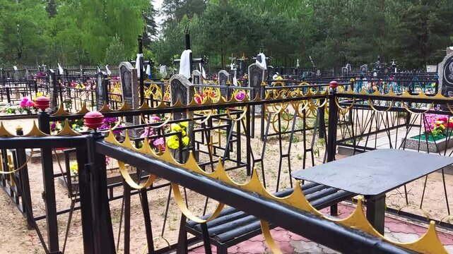 Orthodox cemetery with graves, ornate crosses, artificial flowers, and decorative metal fences. Bench and table on sandy ground. Green trees in background. Scene for Radonitsa holiday.