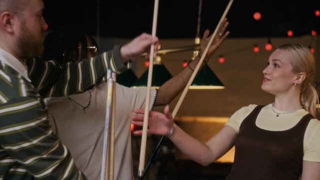 Tilt up shot of three excited young adult friends picking up cues while getting ready to play billiards game at pool club