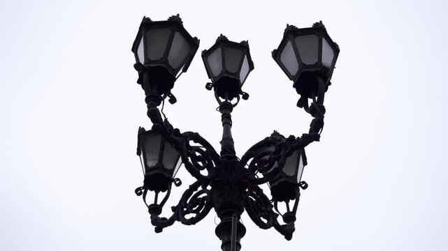 Ornate lamppost silhouette features multiple traditional light fixtures on a decorative wrought iron pole, standing against an overcast sky, providing street lighting ambiance.