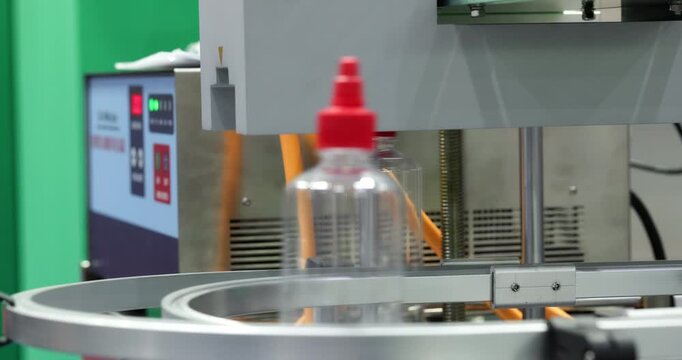 Plastic bottles move along circular conveyor and align under dispensing unit before filling stage. Preparation step in automated bottling and packaging process.